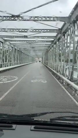 The video shows a view from inside a vehicle driving across a bridge. The bridge is a metal truss bridge with a grey structure. The road is wet, indicating it is raining, and raindrops are visible on the windshield. There are lane markings on the road, including a bicycle lane symbol. Other vehicles are visible on the bridge in the distance. The environment outside the bridge appears to be somewhat urban or suburban, with buildings visible in the distance.