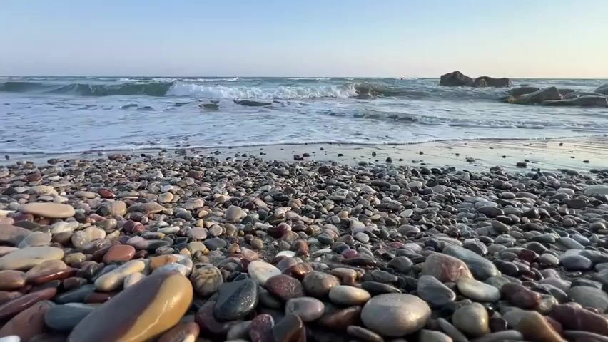 This media file captures a serene scene at a pebble beach in Episkopi, Cyprus. The low-angle shot focuses on the vast expanse of smooth, multi-colored pebbles that make up the foreground. These stones, in shades of grey, brown, pink, and white, are wet and glistening, suggesting the proximity of the sea.

Beyond the pebble shore, the gentle waves of the Mediterranean Sea are rolling in, creating foamy white surf that washes over the stones at the water's edge. The water itself is a clear turquoise, transitioning to a deeper blue towards the horizon. In the distance, larger rock formations rise out of the sea, adding texture to the seascape.

The sky above is a clear, pale blue, indicating fair weather and likely late afternoon or early morning. There are no people or man-made objects visible in the frame, emphasizing the natural beauty and tranquility of the location. The overall mood is one of peacefulness and natural immersion, inviting the viewer to experience the calming rhythm of the waves and the tactile sensation of the pebbles. The lack of visible text further enhances the natural, undisturbed atmosphere.