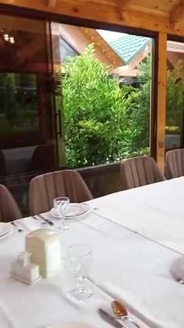 The video pans across a long, white-clothed table set with plates and cutlery, in what appears to be an outdoor dining area with a wooden roof. Large glass windows offer views of lush green trees and foliage. Several chairs with brown upholstery are positioned around the table. A person in a pink garment is briefly visible through the windows in the background. The overall impression is of a restaurant or outdoor event space.