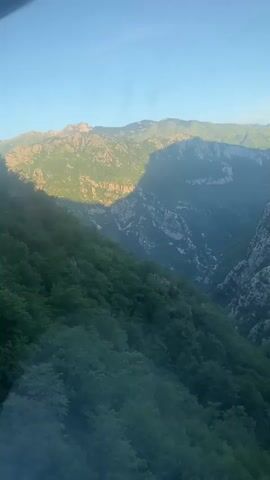 The video shows a panoramic view of a mountain valley. In the foreground, there is a dense forest with green trees. The forest slopes down into a deep canyon, with steep, rocky cliffs on the other side. The mountains in the background are bathed in sunlight, creating a dramatic contrast with the shadows in the valley. The sky is clear and blue. The camera appears to be moving slightly, as if it is on a vehicle or being held by someone walking. There are no people or objects visible in the video. The environment is natural and unspoiled.