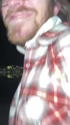 A person wearing a red and white flannel shirt and glasses walks along a sidewalk at night. Buildings and parked cars are visible in the background. The person's face is briefly visible at the beginning of the video. The person seems to be smiling.