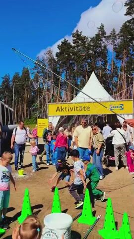 A wide shot captures a lively outdoor scene with a person holding a large bubble wand, creating numerous bubbles that float across the blue sky. Below, a white tent with a yellow banner displaying "Aktivitāte" and an IKEA logo stands prominently. A crowd of people, including adults and children, mill about in front of the tent, some observing the bubbles while others engage in activities. Two children in the foreground are particularly active, with one in a blue shirt and white shorts and another in a green tracksuit. In the background, a dense line of tall pine trees borders the area, and a white traffic sign with a red circle and a white horizontal bar is visible. The bright sunny weather contributes to the vibrant atmosphere of the event.
