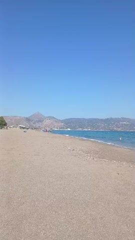 A tranquil beach scene unfolds under a clear blue sky. The foreground is dominated by a wide expanse of light brown sand, leading to the gentle turquoise waters of the sea. In the distance, a hazy coastline stretches horizontally, with rolling hills and some scattered buildings suggesting a populated area. The overall atmosphere is serene and peaceful.

The activity taking place is very minimal, hinting at a relaxed day at the beach. A few distant figures can be seen in the water, either swimming or wading. On the sand, there are a couple of people walking along the shoreline, their presence adding a sense of scale and life to the vastness of the beach. The sunlight suggests it's daytime, likely late morning or afternoon, given the clear sky and bright illumination. The weather appears to be warm and pleasant, ideal for a beach outing.

Notable details include the clear, unbroken blue sky, indicative of fair weather. The gentle waves lapping at the shore create a soothing visual and implied auditory experience. The distant mountains and the strip of settlements on the coast provide subtle location cues, hinting at a Mediterranean setting, possibly the Greek coast as suggested by the location context. The overall mood is one of calm, relaxation, and enjoyment of a natural landscape. No visible text is present in the media file.