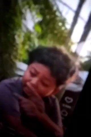 The video shows two young boys outdoors. The first boy, who appears to be in his early to mid-childhood, has dark, curly hair and is wearing a dark t-shirt. He is looking downwards with a thoughtful or sad expression. The second boy, also a child, is partially visible in the foreground, looking to his left. They are in an environment with lush green foliage, including palm trees, and possibly some man-made structures in the background. The lighting suggests it is daytime. The video is low resolution and blurry, making it difficult to discern fine details. There are on-screen overlays indicating social media interaction metrics like likes and comments.