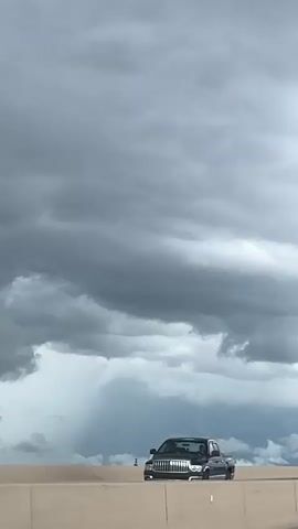 The video shows a view from a vehicle driving on a highway. The sky is overcast with dark, dramatic clouds. In the distance, rain can be seen falling. Several cars are on the road, including a white SUV and a silver sedan. Streetlights are visible along the side of the highway. The camera is positioned to capture the road ahead and the sky above.
