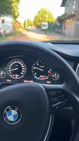 The video shows the interior of a BMW from the driver's perspective. The focus is on the steering wheel, dashboard, and the view through the windshield. The car appears to be stationary on a residential street. The dashboard displays various gauges and information. The steering wheel is black leather with the BMW logo prominently displayed. Through the windshield, we can see a street lined with houses and trees. The lighting indicates it is daytime. There is no visible driver or other occupants in the car. The camera is static, providing a fixed perspective.