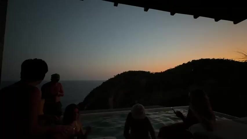From under a roof, a group of people are silhouetted against the backdrop of a twilight sky, a dark green hillside, and the ocean. Five people are near a square pool, possibly a hot tub. One person, seated on the edge, is holding a phone. One is submerged in the water, while the remaining three stand nearby. The sky transitions from dark blue to orange at the horizon, where the sun appears to be setting.