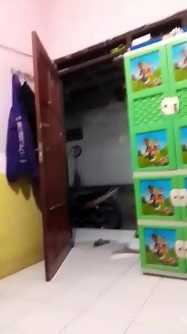 The video shows an interior space that appears to be a workshop or storage area. In the foreground, a stack of colorful folded fabrics is visible on the left. To the right, a bright green plastic storage cabinet with cartoon images is partially visible. A dark-colored motorcycle is parked in the background, partially obscured by an open, dark red door. A sewing machine is set up on a table with a blue surface, and a folding chair is positioned in front of it. Other objects, such as a fan and shelves with various items, are also present. A child with dark hair is briefly visible in the bottom left corner of the frame. The walls are painted in light colors, and the floor appears to be tiled. The overall lighting is dim.