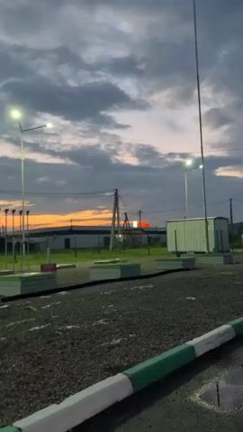 The video begins with a blurred shot of a parking lot at dusk. A streetlamp is visible, along with some buildings in the background. The sky is cloudy with an orange glow from the setting sun. The camera then pans to the left, showing more of the parking lot and a fence. As the video progresses, the camera moves further inside what appears to be a vehicle. The last shot shows a young woman with blonde hair and blue eyes leaning out of the car window, looking at the camera. She has her lips pursed. The lighting is dim, suggesting it is nighttime or very late dusk.