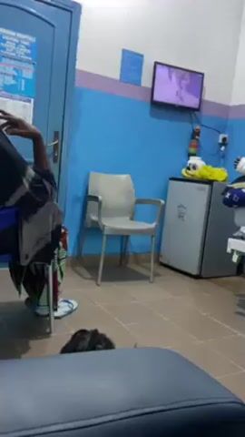 The video shows an indoor scene of what appears to be a waiting room in a hospital or clinic. The walls are painted in blue and white, with a horizontal purple stripe. A woman wearing a patterned dress and slippers is seated in the lower left corner, partially obscured by the camera's perspective. In the center of the frame, there's an empty light grey plastic chair. To the right of the chair, a small silver refrigerator stands against the wall. A television is mounted on the wall above the refrigerator, displaying a show. The floor is tiled in a neutral color. Other objects include a bed with a patient to the right, a sign posted on a door to the left, and various items on top of the refrigerator. The lighting is artificial and appears to be from overhead fixtures.