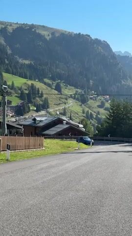 A blue vehicle drives on a paved road in a mountainous region. The road curves to the right. On the left of the road, there is a wooden fence, green grass, and a ski lift. Several buildings with brown roofs are situated on a hillside behind the fence. The hillside is covered with green grass and trees. In the background, there is a large mountain range covered with trees. The sky is clear and blue.