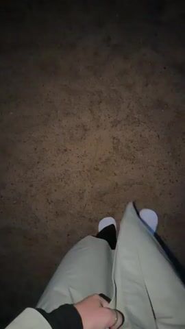 The video shows a first-person perspective of someone walking on a sandy or dirt surface at night. The person is wearing light-colored pants, black leggings or tights, and white socks. The camera is pointed downwards, focusing on the person's legs and feet as they move. The background is dark and appears to be an outdoor environment. The person's hand is visible in the lower left corner, holding onto their pants. The action consists of repetitive forward steps, indicating walking.