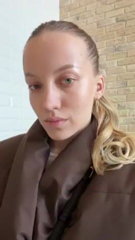 A light-skinned woman is drinking from a white paper cup with a black lid. She is indoors, with a brick wall visible behind her. She is wearing a brown puffy jacket and has her blonde hair pulled back in a ponytail with curls at the end. She has simple gold earrings. She is also wearing a black crossbody bag. After drinking, she looks directly at the camera.