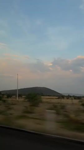 The video shows a landscape as seen from a moving vehicle. In the foreground, there is dry grass and small green trees, with a highway guardrail visible at the bottom of the frame. In the middle ground, there is a hill covered in vegetation. In the background, there is a sky with a mix of blue and gray clouds, some edged with a faint pinkish hue, suggesting either sunrise or sunset. A utility pole and wires cut across the scene. The camera is moving, blurring the foreground details.