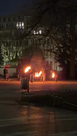 The video shows several individuals performing a fire dancing routine in a park at night. There are five people, including a woman wearing a crop top and camouflage pants spinning two objects that emit fire. A man to her right is using a chain to spin a flaming object. The other members of the group are also handling flaming objects. The performance takes place on an asphalt surface. A trash can and bicycle rack are visible in the foreground. In the background, there is a building with lights on. Bare trees and a dark sky complete the scene.