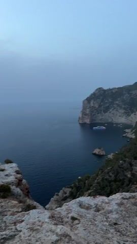 A woman with long brown hair walks along a rocky cliff overlooking a vast body of dark blue water. She is wearing a light blue, short, sparkly dress and black sandals. In the distance, a large boat is visible on the water. The sky is a muted gray with some lighter patches, suggesting either dawn or dusk. The terrain is rugged and uneven, with sparse green vegetation clinging to the rocks. The woman is walking away from the camera, her figure silhouetted against the expansive seascape.