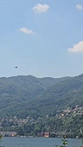 A helicopter flies across a bright blue sky with scattered white clouds. Below the helicopter, a lush green mountainside is dotted with a village of colorful houses, appearing to cling to the steep slope. The village extends down to the edge of a large, calm body of water, which occupies the bottom portion of the frame. Along the waterfront, several buildings are visible, some appearing larger and more elaborate than the village houses. The scene suggests a picturesque, possibly Mediterranean, location on a sunny day. The overall impression is one of natural beauty combined with human settlement, with the helicopter adding a dynamic element to the landscape. There is no visible text in the media.