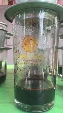 A close-up shot shows coffee dripping into a glass. The camera then pans to reveal several other glasses filled with coffee, followed by a person wearing a red hat and a face mask sitting on a chair and eating. The scene then shifts to show a street vendor's stall with a pile of coconuts and pineapples, and a truck in the background.