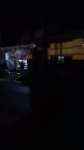 The video begins with a dark, grainy view of a street at night. A building with a red sign featuring images of food is visible on the left, possibly a restaurant or food stall. Several figures are dimly illuminated in front of the establishment, and what appears to be a motorcycle is parked nearby. The camera then pans to the right, revealing a covered vehicle parked under a structure. The scene transitions to a completely dark frame for a moment, followed by intermittent flashes of light and glimpses of illuminated structures in the distance. The video concludes with a very dark frame, showing faint, distant lights against a black background.