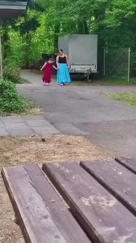 A woman and a young girl walk hand-in-hand down a paved path surrounded by trees and grass. The woman wears a black tank top and a long blue skirt. The girl wears a red dress. A light-colored trailer with a gray tarp is parked on the right side of the path behind them. A dark-colored dumpster is visible on the left side of the path. The foreground shows a close-up of a wooden picnic table.