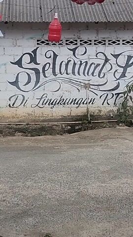 The video captures a street scene in Tegal Rejo, Indonesia. The camera is oriented vertically, presenting a slightly tilted perspective of the environment.

The first few seconds show a wall with large, stylized black lettering that reads "JAMAL BARBER LINGKUNGAN RT 3". To the left of the wall is a corrugated metal roof structure, and a bright red plastic water dispenser is visible. A small plant grows out of the wall near the text.

The scene then shifts to a black vehicle, possibly a small truck or utility vehicle, with a metal rack on top. The front tire is visible, and the driver's side door is slightly ajar. A person's reflection can be seen in the window. Beyond the vehicle, there's a glimpse of a green motorcycle and more of the tiled or concrete ground.

Further into the video, the view moves to an area that appears to be an outdoor workspace or storage area. Concrete blocks with red and white striped markings are present, along with a yellow cloth and a red hose. A green bush with leaves is also visible.

The final segment shows a collection of items on the ground, including a red compressor tank, a black metal structure, a yellow and black object that might be part of a tool, and a coiled hose. Several plastic bottles, some containing liquid, are also scattered around, along with some white plastic cups. A red cap or helmet can be seen in the lower right corner.

The overall impression is of a utilitarian, somewhat cluttered outdoor area, likely in a residential or small business setting. The lighting suggests daytime, but the weather is not definitively indicated. There are no overt signs of emotional expression or a specific event beyond the general activity of the location.