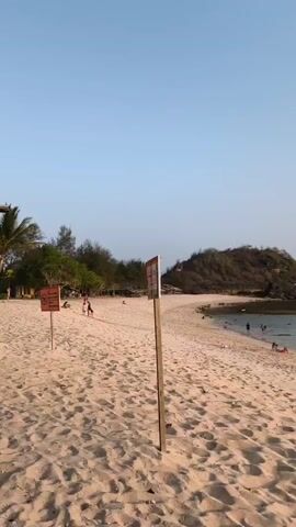 The video shows a sandy beach with a clear sky. There are two orange signs with black symbols and text on wooden posts in the foreground. The sign on the left has a symbol of a dog with a line through it and text that reads "DILARANG BERENANG" and "DON'T SWIMMING". The sign on the right has a red symbol with text that is difficult to read. In the background, there are trees, some small structures that appear to be huts or stalls, and a few people walking and sitting on the beach. The water is visible in the distance. The overall scene depicts a typical beach environment with some regulations in place.