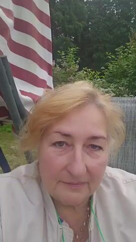 A close-up, slightly shaky video features a woman with blonde hair and fair skin, looking directly at the camera. She is wearing a light-colored jacket with a prominent zipper and a necklace. A pair of glasses is hanging from a green cord around her neck. Behind her, a striped fabric, red and white, is visible, along with greenery and trees, suggesting an outdoor setting. The lighting suggests it is daytime, likely overcast or in shade, as there are no harsh shadows. The woman's expression is neutral, perhaps with a hint of a smile. The background is somewhat blurred, with a focus on the woman's face. The overall impression is of a casual, personal recording, possibly a selfie taken in a garden or park. There is no visible text in the video.