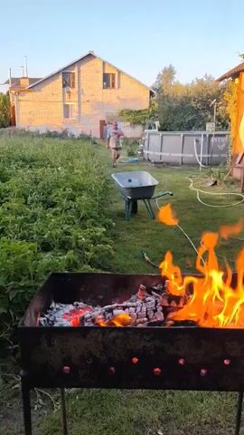 The video shows a backyard scene with a barbecue grill in the foreground, flames and glowing embers visible within. Beyond the grill, a person is walking away from the camera towards a residential building. A wheelbarrow is positioned on the grass between the grill and the person. In the background, there's a large inflatable swimming pool and a wooden structure. The environment appears to be a garden with lush green plants. The lighting suggests it is either early morning or late afternoon.