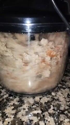 A video of a kitchen countertop shows a blender or food processor with a clear bowl and a black lid, on a speckled granite surface. Inside the bowl, light brown, fibrous material is being chopped by a spinning blade, creating a fine texture. The camera angle shifts slightly, showing more of the black lid and a cord plugged into an unseen power source.