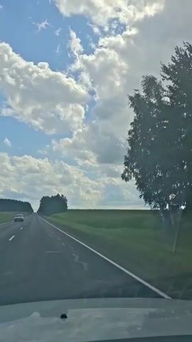 The video captures a first-person perspective from inside a vehicle traveling on a paved road. The road stretches ahead into the distance, bordered by a lush green field on the right and trees on the left. The sky above is a vibrant blue, dotted with fluffy white clouds. Another vehicle is visible in the distance on the left side of the road. The sun appears to be shining, casting light and shadows. The overall impression is that of a scenic drive through a rural landscape.