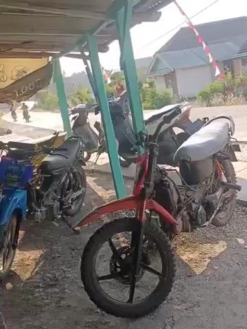 The video shows several motorcycles parked under a structure with a corrugated metal roof. The motorcycles are varied in color and style, with one prominently featuring a red frame and off-road tires. The ground is covered in gravel. In the background, there are houses and trees, suggesting a rural or suburban setting. Sunlight indicates it is daytime. A person is visible in the distance near one of the houses. The overall impression is a casual outdoor scene with parked vehicles.