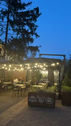 The video shows an outdoor seating area of a restaurant or cafe at dusk. A string of lights is visible overhead. Several people are seated at tables, some eating and others talking. A few people are walking around the area. The ground is paved with bricks. There are trees and planters with greenery. The sky is a deep blue, indicating twilight. The scene is peaceful and relaxed.