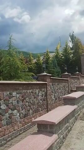 The video shows a view from a stone wall with a pink granite top to a large building, likely a hotel or resort, with multiple floors and balconies. The building has a rustic architectural style with wood and stone accents. Evergreen trees surround the building, and a red car is parked in front. The sky is cloudy with some blue patches, and a green, tree-covered hill is visible in the background. The stone wall itself is constructed with various colored stones, including grey, pink, and brown. There are decorative lamps on stone posts that line the top of the wall.