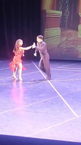 The video shows a male and female dancer performing on a stage. The female dancer wears a red, short, fringed dress with a high leg slit and heels. The male dancer wears a black long-sleeved shirt and black pants. They are holding hands and moving in a dance routine, with the female dancer performing a high leg kick. The stage floor is purple with white lines and there is a black backdrop behind the dancers. A blurry image of a scenic backdrop is visible in the upper right corner of the frame. The lighting is dim with spotlights illuminating the dancers.