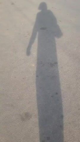 The video features a person's long shadow cast on an asphalt road in Barden Yana, Nigeria. The shadow, tall and elongated, suggests the sun is low in the sky, indicating either early morning or late afternoon. The person, whose form is only visible as a shadow, momentarily raises a hand to their head. A red car then approaches from the distance, driving directly towards the camera. The front of the car, including its headlights and grille, becomes clearly visible as it draws closer, while the person's shadow remains prominent in the foreground. The road appears dry and clear.