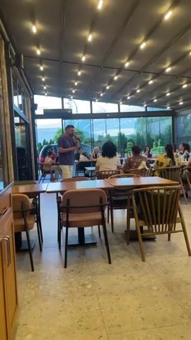 A man stands holding a microphone and speaking to a group of people seated at tables in what appears to be a restaurant or cafe. He is wearing a patterned shirt and white pants. The people at the tables are mostly women, and some are looking towards the man. The environment has large windows and overhead lights. The man is in the foreground, and the seated patrons are in the mid-ground.