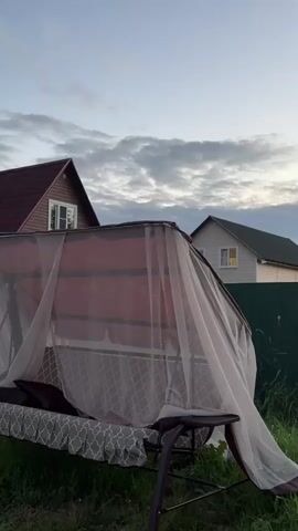 A swing set with a mosquito net is in the foreground. In the background, there are two houses, a dark green fence, and trees. The sky is cloudy. The video pans from left to right.