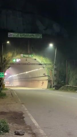 The video shows the entrance of a tunnel labeled "TUNNEL NO. 5" with the National Highways Authority of India welcoming visitors. The tunnel is brightly lit from within, creating a strong contrast with the dark exterior. A road leads up to the tunnel, and a few vehicles, including a truck and a bus, are seen either approaching or entering the tunnel. The entrance is adorned with traffic lights and signage. The overall scene suggests a nighttime view of a major road infrastructure project.
