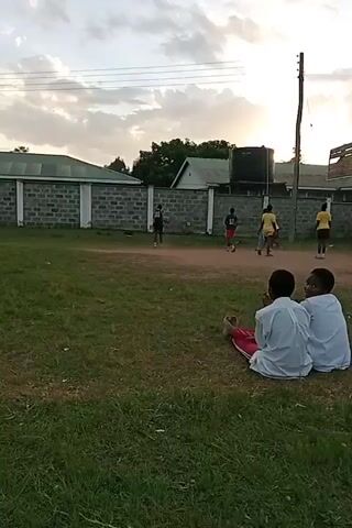 Here's a description of the video:

The video shows a group of children playing basketball on a dirt court. The court is located in an open field next to a low brick wall and some buildings. Two children are sitting on the grass in the foreground, watching the game. They are both wearing light-colored shirts, and one is wearing red pants. 

The basketball players are wearing a mix of clothing, including yellow jerseys and black shorts. The sun is setting in the background, creating a warm glow in the sky.