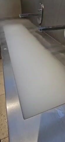The video shows a close-up, angled view of a large, stainless steel sink. Two faucets are visible at the top of the sink, and a dense cloud of white vapor emanates from the sink basin. The vapor is thick and obscures the bottom of the sink and the water flow from the faucets. The vapor is actively being produced, swirling and dissipating slightly at the edges. The environment appears to be indoors, possibly a public restroom or a commercial kitchen due to the sink and faucet style.