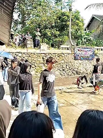 The video captures a daytime outdoor scene featuring several people walking through what appears to be a small town or village. The ground is wet, possibly due to recent rain. A man in a black t-shirt and blue jeans walks in the foreground holding a pair of white sneakers. Next to him, a woman in a black hijab and denim jacket also walks through the scene. Further into the shot, another man in a grey hooded sweatshirt and jeans is walking. Behind them, other individuals stand along a stone wall, observing the activity. Some are holding umbrellas. A few people are wearing traditional patterned clothing and appear to be part of a performance. A banner is visible behind the stone wall. In the background, there are houses with corrugated roofs. The environment suggests a rural setting, and the presence of multiple people indicates an event or gathering is taking place.