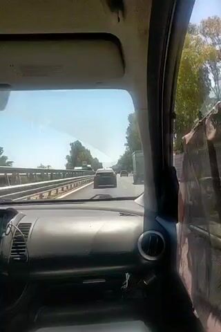 The video shows the interior of a car from the driver's perspective, looking out through the windshield. The car is on a highway with multiple lanes of traffic moving in the same direction. There are other vehicles on the road ahead. The landscape outside the car consists of trees and vegetation on either side of the highway. The sky is clear and blue, indicating a sunny day. The dashboard and part of the car's interior are visible in the foreground. The video appears to be a recording of a car journey.