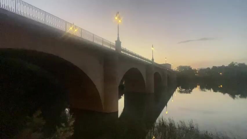 This is a video clip showing a stone bridge with multiple arches spanning a calm river. The bridge has a railing along its top and several ornate street lamps that are illuminated, casting a warm glow. The river reflects the bridge and the lights, creating a symmetrical and serene image. The sky is a gradient of dusk colors, transitioning from a pale yellow near the horizon to a soft purple and blue overhead. Silhouetted trees line the far bank of the river, and a few distant lights suggest a town or settlement. The overall mood is peaceful and tranquil, evoking the quiet beauty of early morning or late evening. The time of day is clearly twilight. There are no people visible in the clip. The location is identified as Logroño, Spain, which is known for its historical bridges over the Ebro River.