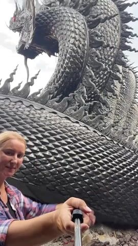 A woman stands in front of a large, dark, dragon-like sculpture. She is wearing a plaid shirt and holding a selfie stick. Her left arm is extended, and her hand is pointing toward the sculpture. The sculpture is intricately detailed, with scales and sharp protrusions. The background shows a cloudy sky.