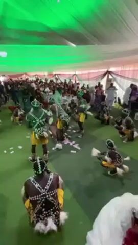 The video shows a group of dancers performing in what appears to be a large tent or indoor event space. The dancers, who are mostly shirtless and painted with green and white body paint, are wearing yellow shorts and beaded headdresses. Some are playing drums while others dance. Scattered on the green floor are pink bills, resembling money. An audience of people is seated around tables in the background. The tent is decorated with white draping and a green light shines down from the ceiling. The camera angle is relatively high, providing an overhead view of the performance area.