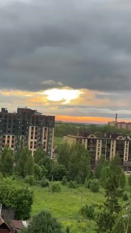 The video shows a panoramic view of a city skyline during a sunset. The sky is filled with dark, heavy clouds, with a bright, golden sun breaking through the clouds in the distance. The cityscape is dominated by tall, modern apartment buildings. In the foreground, lush green trees and grass suggest a natural environment encroaching on the urban landscape. The overall atmosphere is dramatic and somewhat moody due to the interplay of dark clouds and bright sunlight. No people are visible in the video.
