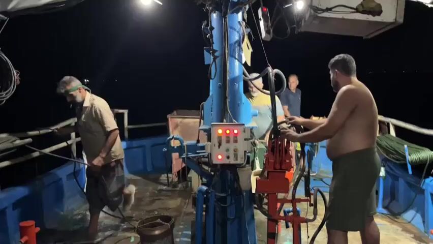 The video captures a nighttime scene on the deck of a fishing boat. Four men are visible. On the left, a man with a beard and a grey shirt is holding a rope and smoking a cigarette. In the center, two men are standing near a control panel with red and green lights. The man on the right of the panel, who is bare-chested and wearing a green wrap, is operating a red lever. Another man in a blue shirt stands behind them. The deck is cluttered with ropes and equipment. The background is dark, suggesting it is open water at night.