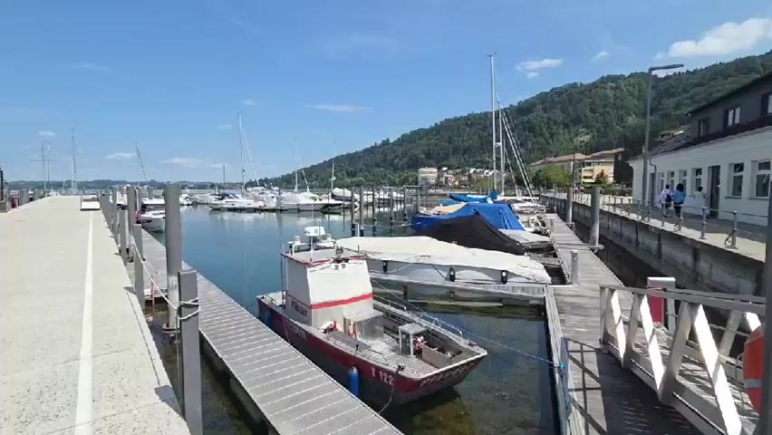 A wide shot of a marina on a sunny day. Numerous sailboats and smaller boats are docked in the calm water. A paved walkway runs along the left side of the frame, with metal posts and railings. A building with a white facade and a dark roof is visible on the right. A gently sloping, tree-covered hill rises behind the marina. Several people can be seen walking on the walkway near the building on the right. The sky is clear and blue with a few scattered clouds. The water reflects the blue sky and the boats.