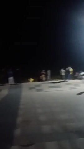 The video captures a nighttime scene in Bagerhat, Bangladesh, likely near a religious site given the mention of "khankajan mazare" (Khankahan's shrine) in the audio. The setting appears to be an outdoor public space, possibly a courtyard or walkway, illuminated by artificial lights, creating a somewhat hazy and atmospheric effect.

Several people are visible, though their features are indistinct due to the low light and motion blur. They are seen walking, standing, and in some instances, gathered in small groups. The activity appears to be casual, with people moving about in the evening. One person is heard speaking in Bengali.

Notable details include the bright overhead lights that cast long shadows, indicative of the nighttime setting. The overall mood seems calm and perhaps a bit solemn, consistent with a visit to a shrine. There are no discernible weather cues, but the darkness suggests it's after sunset.

The audio contains Bengali speech, with the phrase "Bagerhat Khankajan mazare" being the most clearly discernible, indicating the general location. No other visible text is present in the video frames. The subjects are primarily people moving through the dimly lit environment.