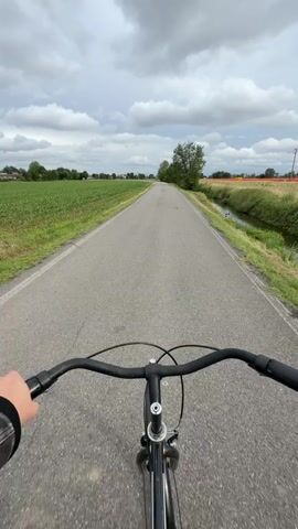 A first-person perspective video shows a bicycle ride on a narrow asphalt road flanked by fields. The fields on the left are green and appear to be planted with crops, while the fields on the right are partially yellowed and bordered by a small stream. The sky is overcast with heavy clouds. The bicycle has a black frame and handlebars. A person's hand wearing a black jacket sleeve is visible holding the handlebar. The road stretches straight ahead, disappearing into the distance.